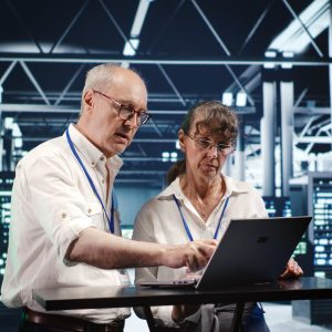 Senescent colleagues looking around high tech data center, using laptop to crosscheck disaster recovery plan and assess server machines in need of replacement, preventing glitches